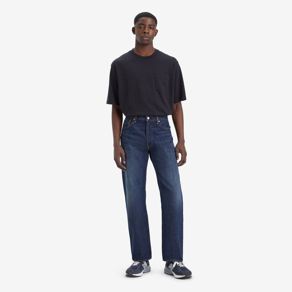 A person stands against a white background in a black t-shirt and Levis 555 Relaxed Straight jeans made from non-stretch denim, complemented by stylish, comfortable sneakers.