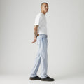 A person in a white t-shirt, 555 Relaxed Straight vintage-inspired jeans by Levis, and black shoes stands against a plain background, looking sideways with hands behind their back.