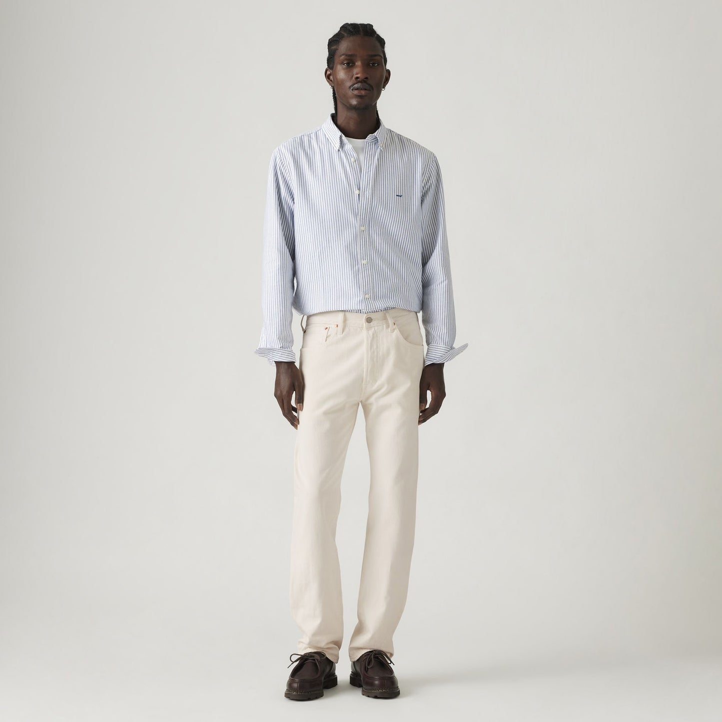 A man stands against a plain backdrop, wearing Levis 501 Original jeans in Rent Free LTWT and a light blue striped shirt, paired with dark shoes.