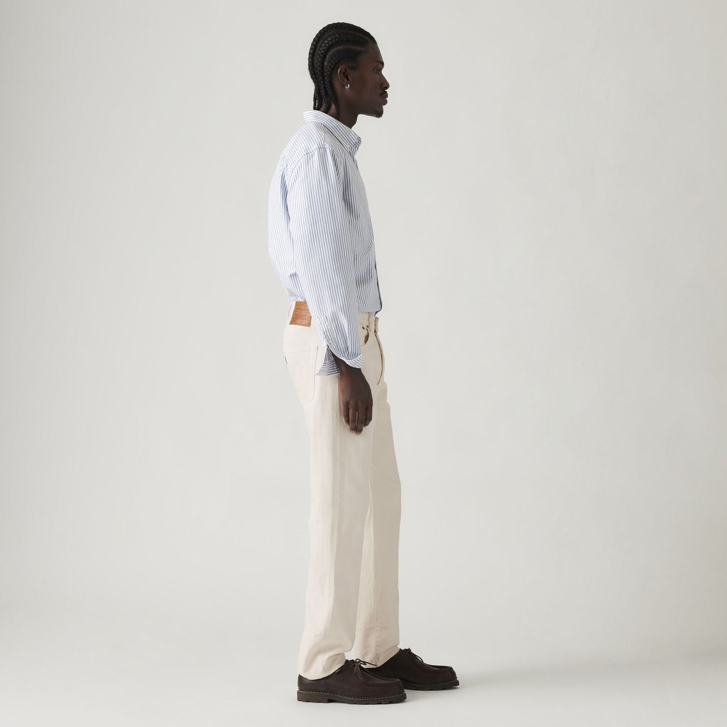 Wearing a light blue Levis Rent Free LTWT shirt, beige 501 Original pants, and brown shoes, the person stands in profile against a plain white background, exuding a relaxed vibe.