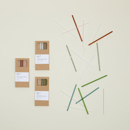 Hawkins New York's Essential Glass Straws - Set of 6, made from durable borosilicate glass, are arranged on a light surface. Three cardboard packages and a cleaning brush lie nearby, highlighting their eco-friendly appeal.