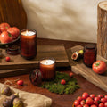 The Amelie - Tomato & Vine 7oz Candle by La Jolie Muse, crafted from natural soy wax and housed in a ribbed glass holder, is complemented by tomatoes, figs, and decorative greenery on wooden boards. A burlap sack and a tree stump add to the rustic backdrop.