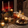 A table set with lit Amelie Linen Cotton Oasis 7oz candles by La Jolie Muse, ribbed glassware, and cutlery. A fireplace in the background enhances the warm ambiance, with a hint of white jasmine wafting through the air.