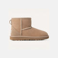 A K Classic Mini II boot by Ugg, featuring tan suede, a flat rubber sole, cushioned outsole, side stitching, and a heel pull tab, is centered against a plain white background.