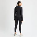 A person with long braided hair in a black Rohnisch Legacy Full Zip outfit and white sneakers stands facing away against a white background. The ensemble, showcasing the Röhnisch logo, is made from recycled polyester, embodying style and sustainability.