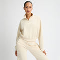 A person models Rohnisch's Soft Sweat Half Zip, featuring a cream hue, long sleeves, and a cropped fit. Paired with matching pants against a white backdrop, the look is stylish and comfy thanks to its Terry fabric.