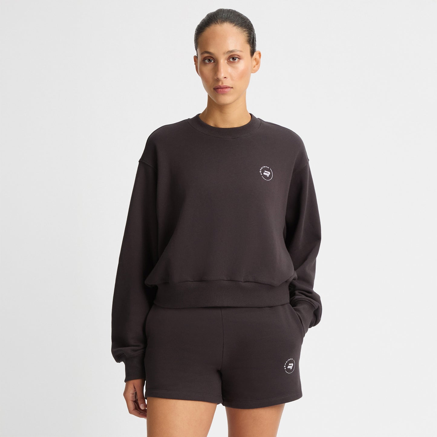 A person stands against a white backdrop, wearing matching dark Rohnisch Soft Sweatshirt and shorts. The cropped top subtly showcases the Röhnisch logo embroidery, crafted from recycled fabric for an eco-friendly touch.