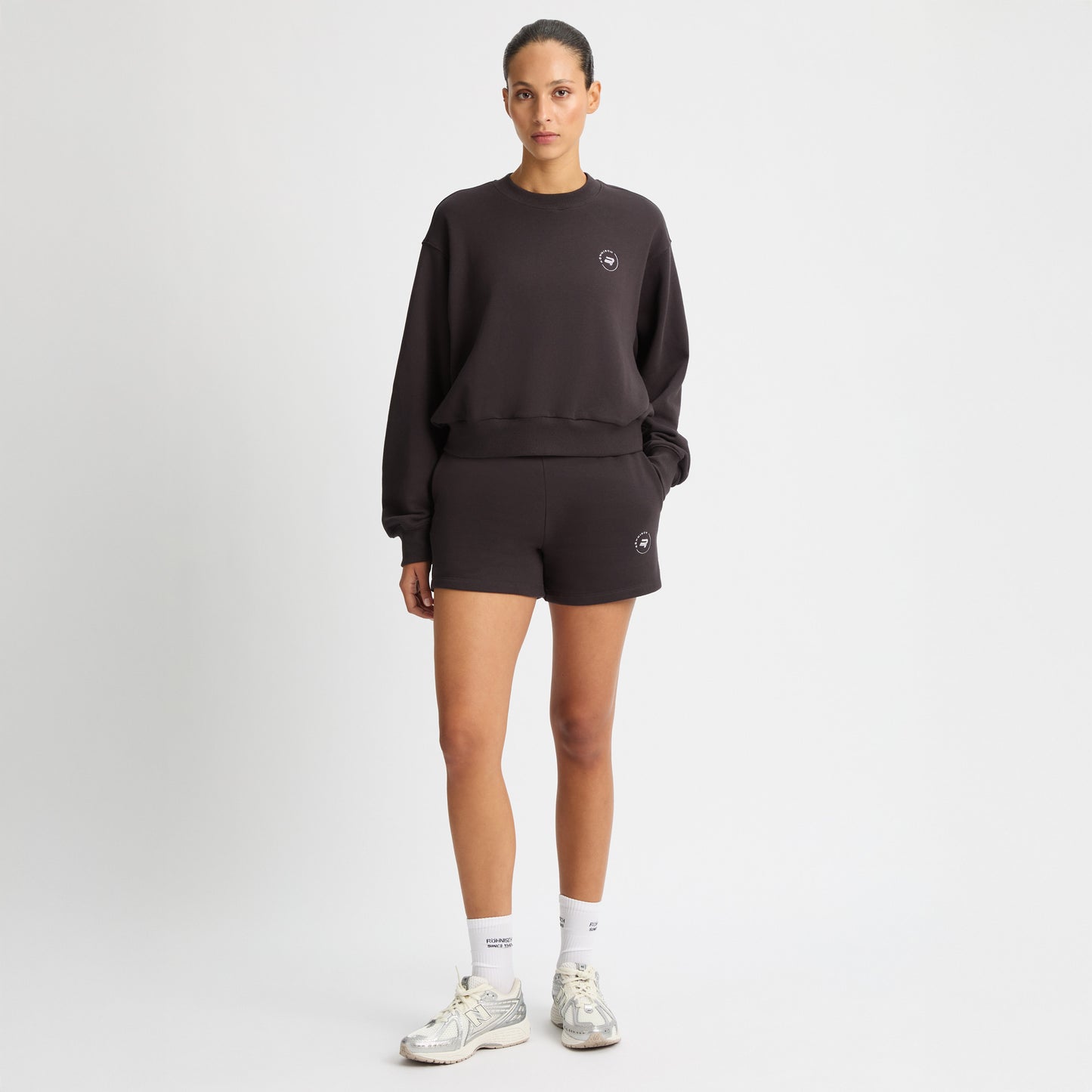 A person wears the Rohnisch Soft Sweatshirt, black and cropped with logo embroidery, paired with shorts, white socks, and silver sneakers against a plain background.