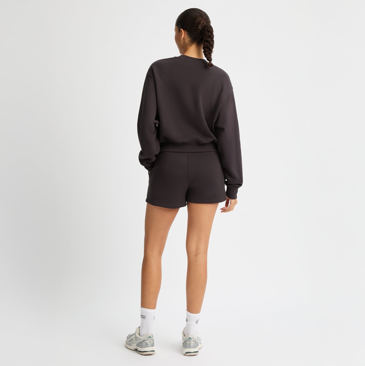 Someone in a dark Rohnisch Soft Sweatshirt and shorts with logo embroidery stands back turned, wearing white socks and gray sneakers. The recycled sweatshirt fabric contrasts against the plain white background, creating a modern, eco-friendly look.