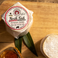 Round of North Fork Whiskey Washed Munster artisan cheese from Redhead Creamery on a wooden board, accompanied by a glass of whiskey from Panther Distillery and green leaves. The cheese is wrapped in paper with a labeled red and white sticker on top.