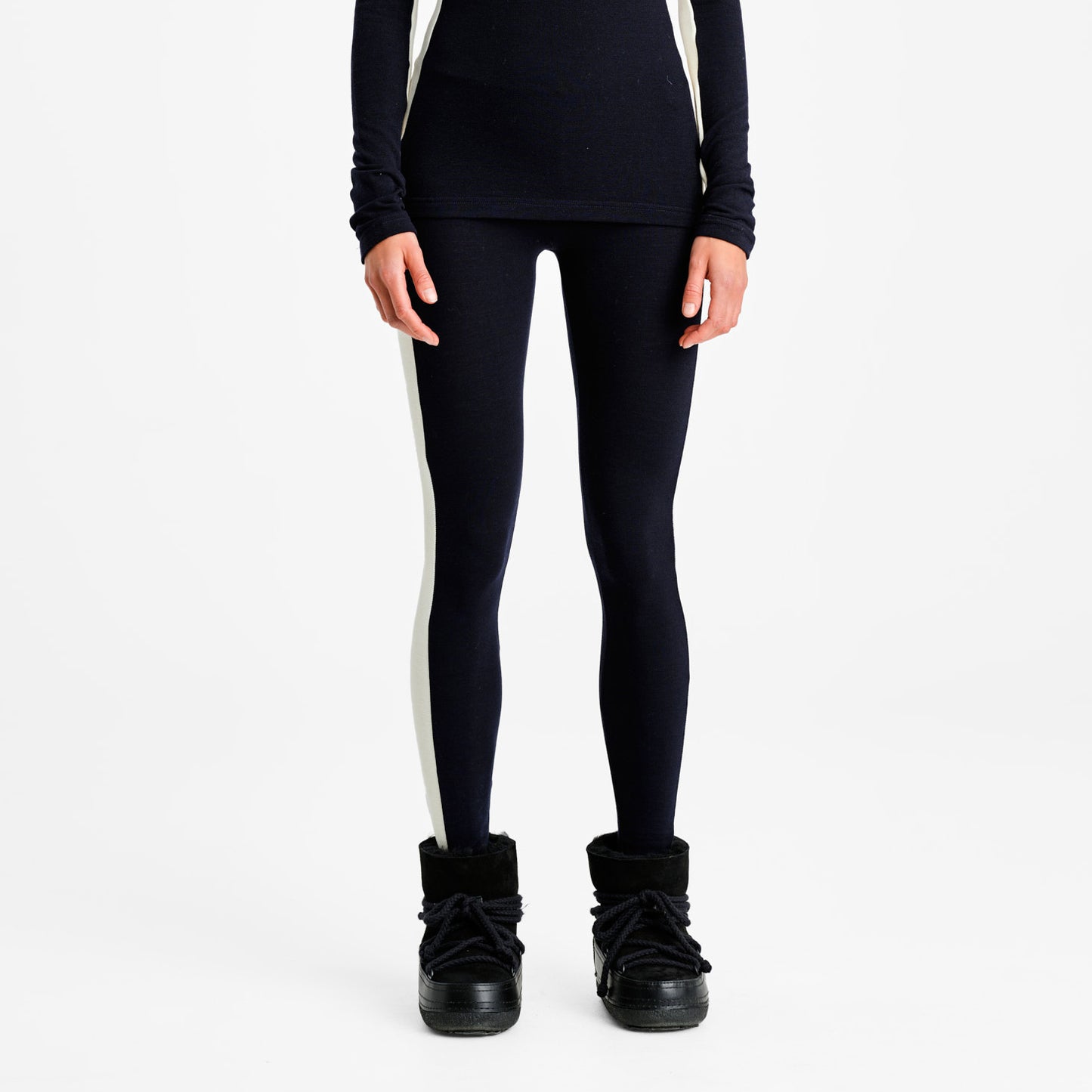 A person wears We Norwegians' W Voss Leggings, featuring black with white side stripes, made from soft Merino wool, paired with black boots, standing against a white background.