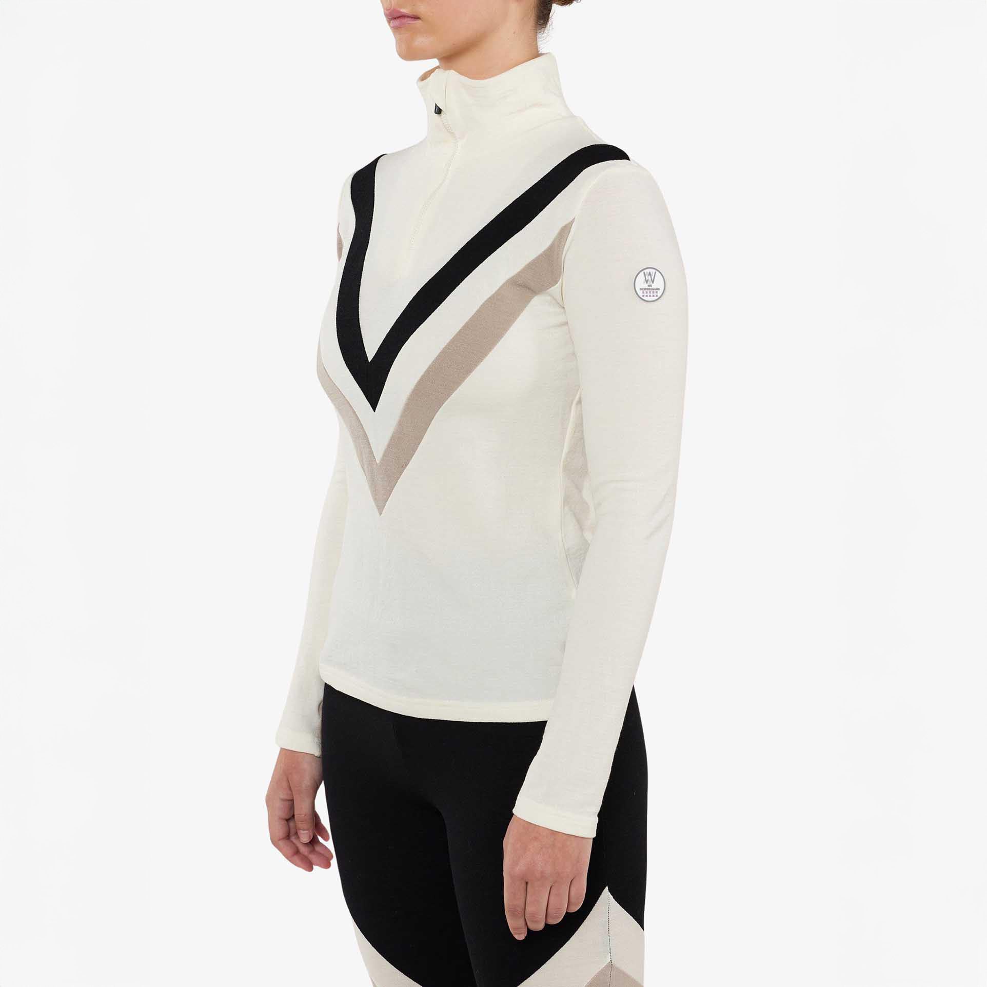 The person is elegantly dressed in the W Voss Chevron Zip-Up by We Norwegians, a long-sleeve, high-neck top crafted from soft merino wool with a bold V-pattern design in black and beige.