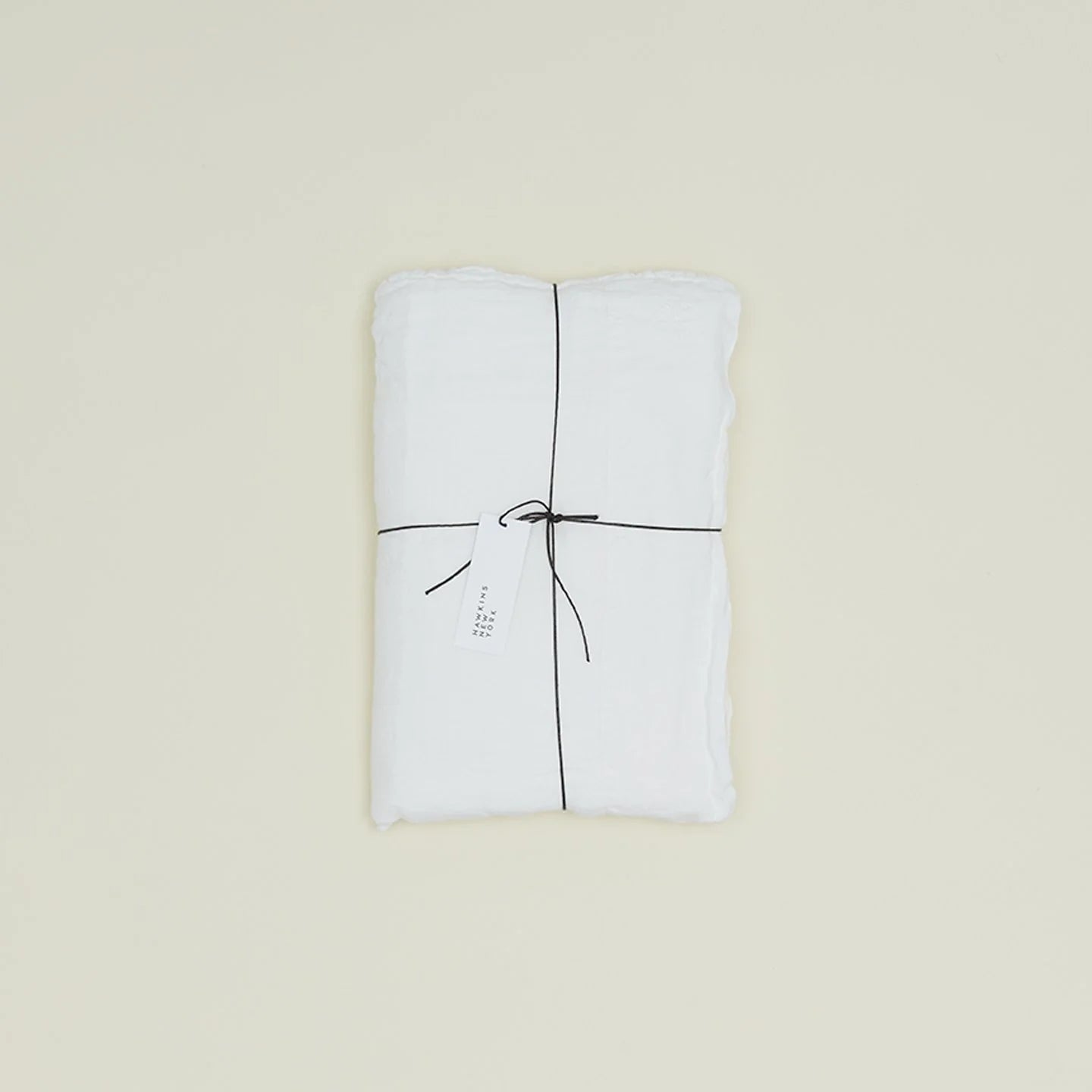 The Simple Linen Fitted Sheet - Queen by Hawkins New York, neatly folded and tied with black string featuring a white tag, graces a plain cream background.