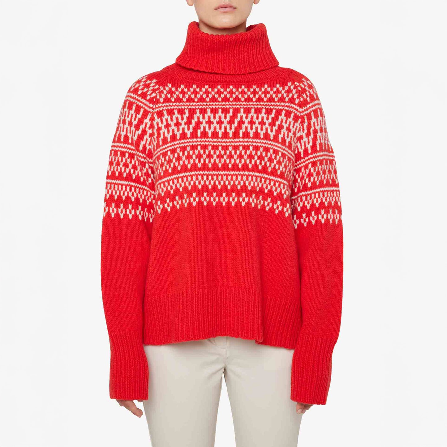 A person wearing a red W Setesdal Sweater by We Norwegians, made of extra fine merino wool with white geometric patterns, paired with light-colored pants against a plain background.