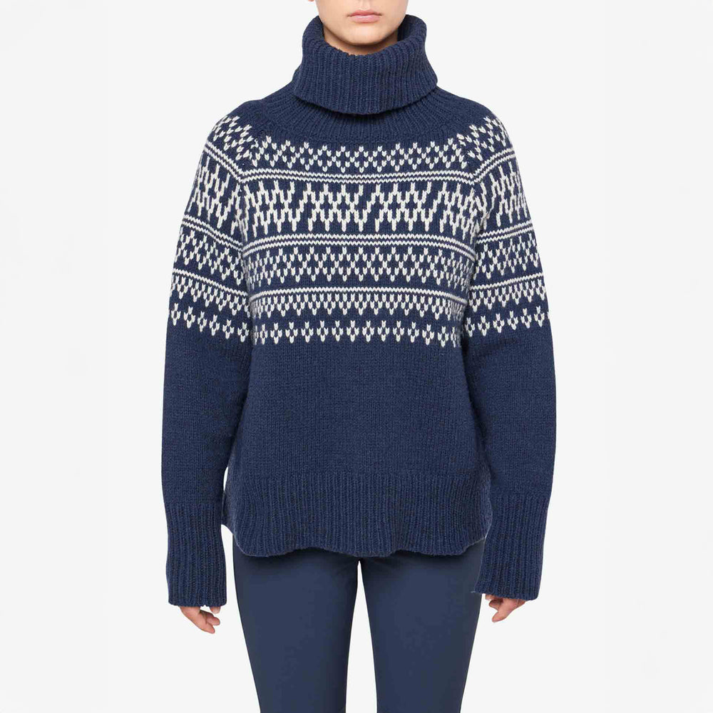 A person wears a W Setesdal Sweater by We Norwegians, featuring a blue and white pattern, paired with navy pants against a plain background.