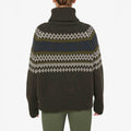 The person is wearing a W Setesdal Sweater by We Norwegians, a dark green turtleneck made of extra fine Merino wool with white and blue patterns, elegantly paired with green pants.