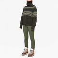 A person in a W Setesdal Sweater by We Norwegians, green pants, and brown boots stands against a white background.