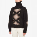 A person is elegantly dressed in a We Norwegians W Hovden Sweater, featuring beige geometric patterns on black lambs wool, designed for women, perfectly paired with crisp white pants.