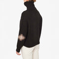 A person wearing the W Hovden Sweater by We Norwegians, a dark lambs wool turtleneck with patterned sleeves, shown from the back, paired with light-colored women's pants.