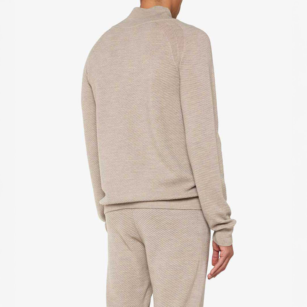 A person models the men's We Norwegians M Geilo 1/4 Zip set, featuring a beige, textured sweater and pants made from luxurious Merino wool, epitomizing elegance against a plain backdrop.