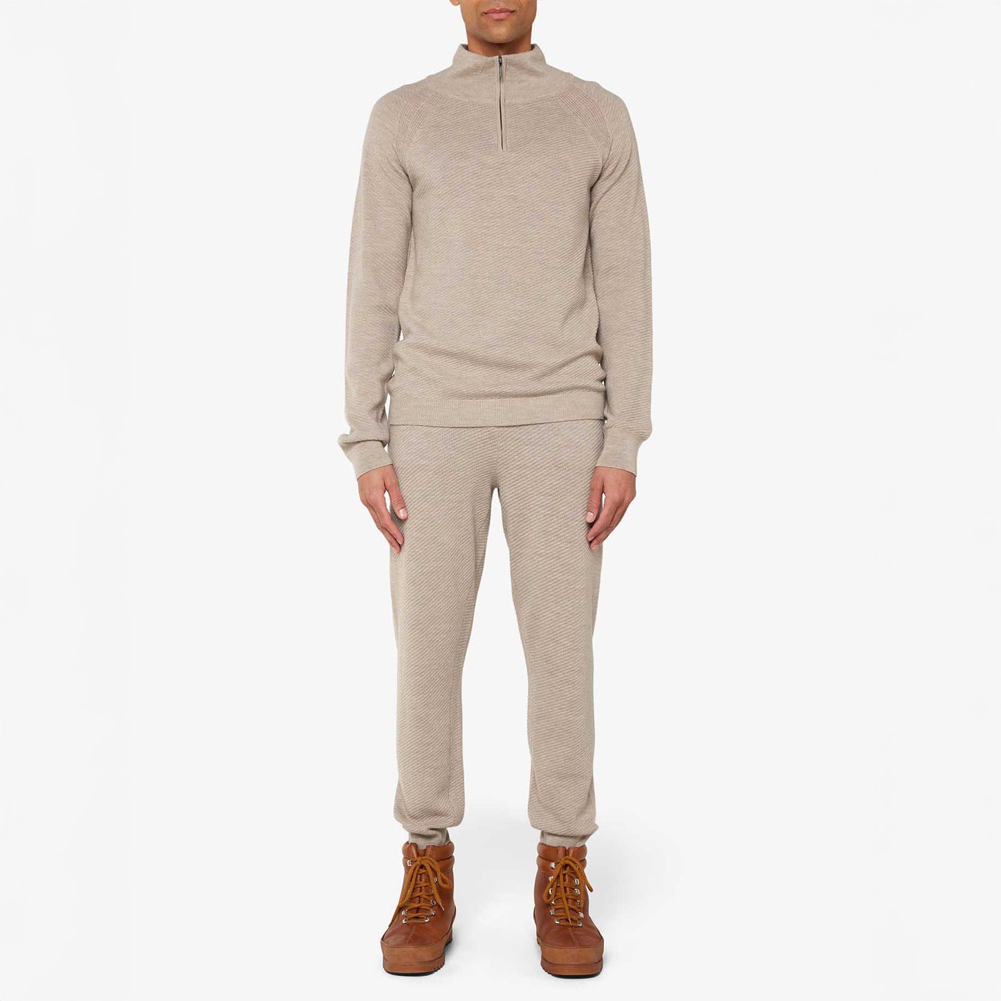 A person dressed in a beige We Norwegians M Geilo 1/4 Zip sweater and matching pants with brown boots stands against a plain background.