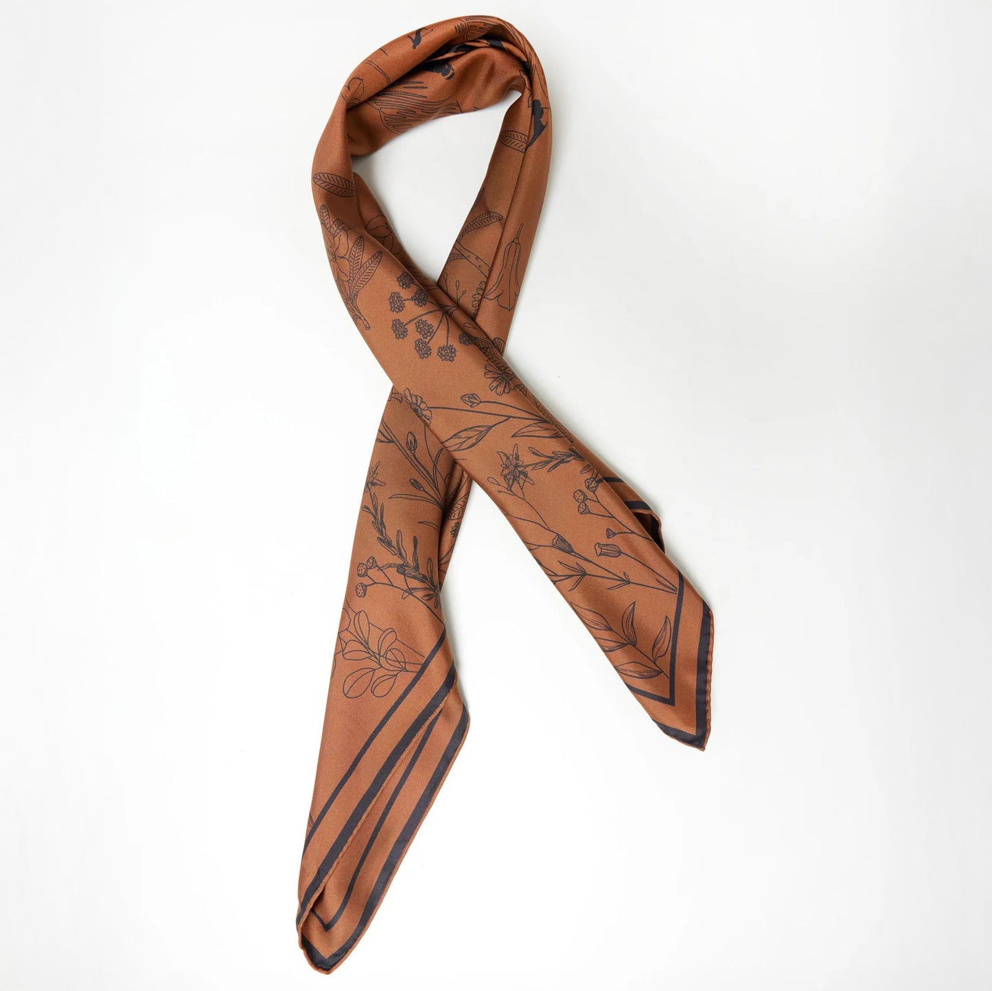 The Alps Silk Scarf by We Norwegians, crafted from pure silk with a brown pattern and black floral designs, is elegantly looped on a light surface, enhancing any ski outfit.