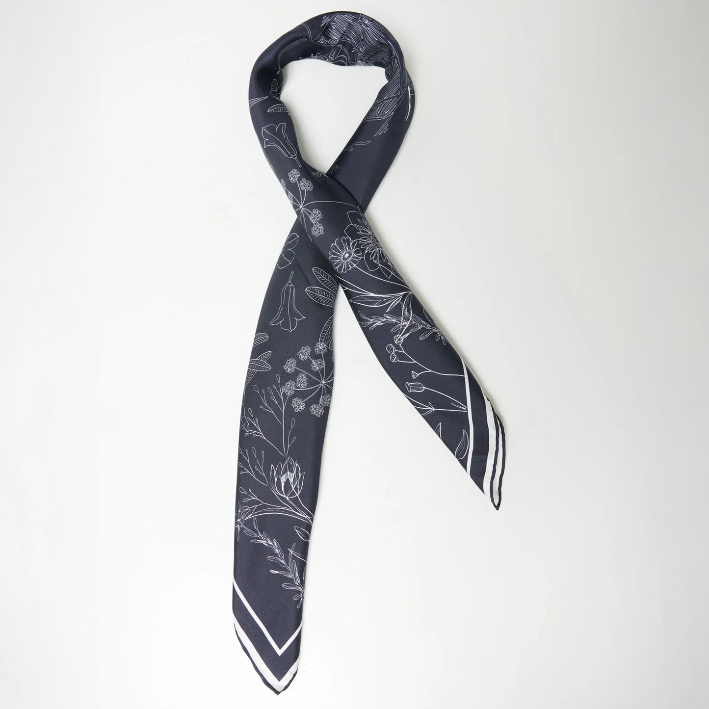 The We Norwegians Alps Silk Scarf, featuring a white floral pattern on black silk, lies flat on a plain white background, adding elegance to any ski outfit.