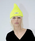 A blond-haired person wears a vibrant Fluo Yellow Logo Beanie by Le Bonnet, celebrated for its durable breathability, paired with a black shirt against a plain background.