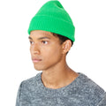 A person wearing a vibrant green Le Bonnet beanie and a gray sweater glances to the side against a white background.