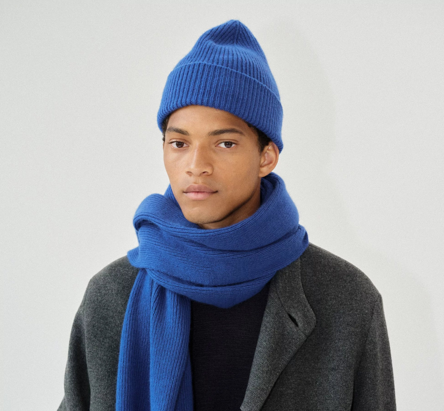 A person in a blue knitted lambswool Le Bonnet hat and the Marin scarf pairs them elegantly with a gray coat against a plain background.