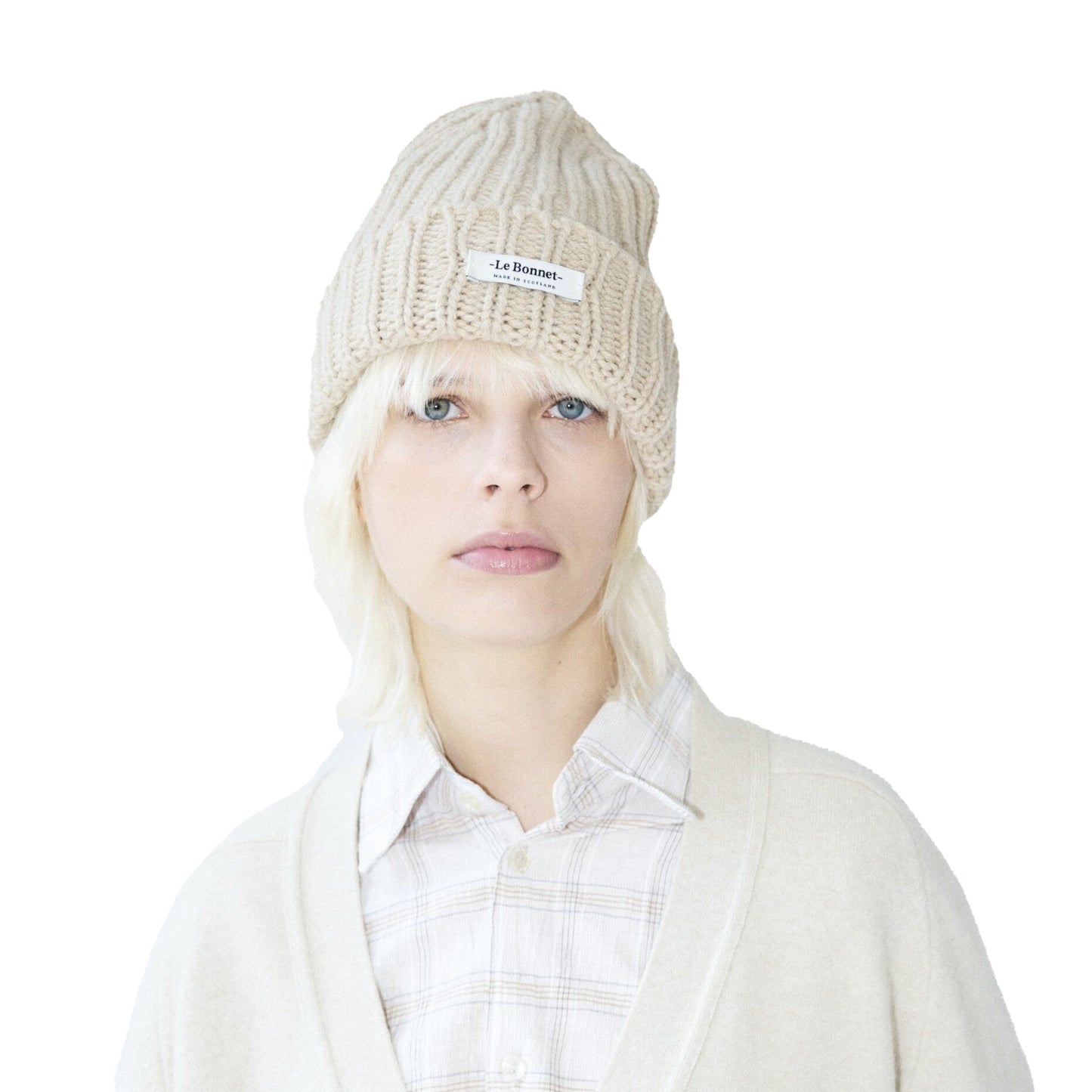 A blonde person models a beige Le Bonnet Logo Beanie and a cream cardigan over a plaid shirt against a white backdrop, highlighting a chic mix of durable and breathable fashion.