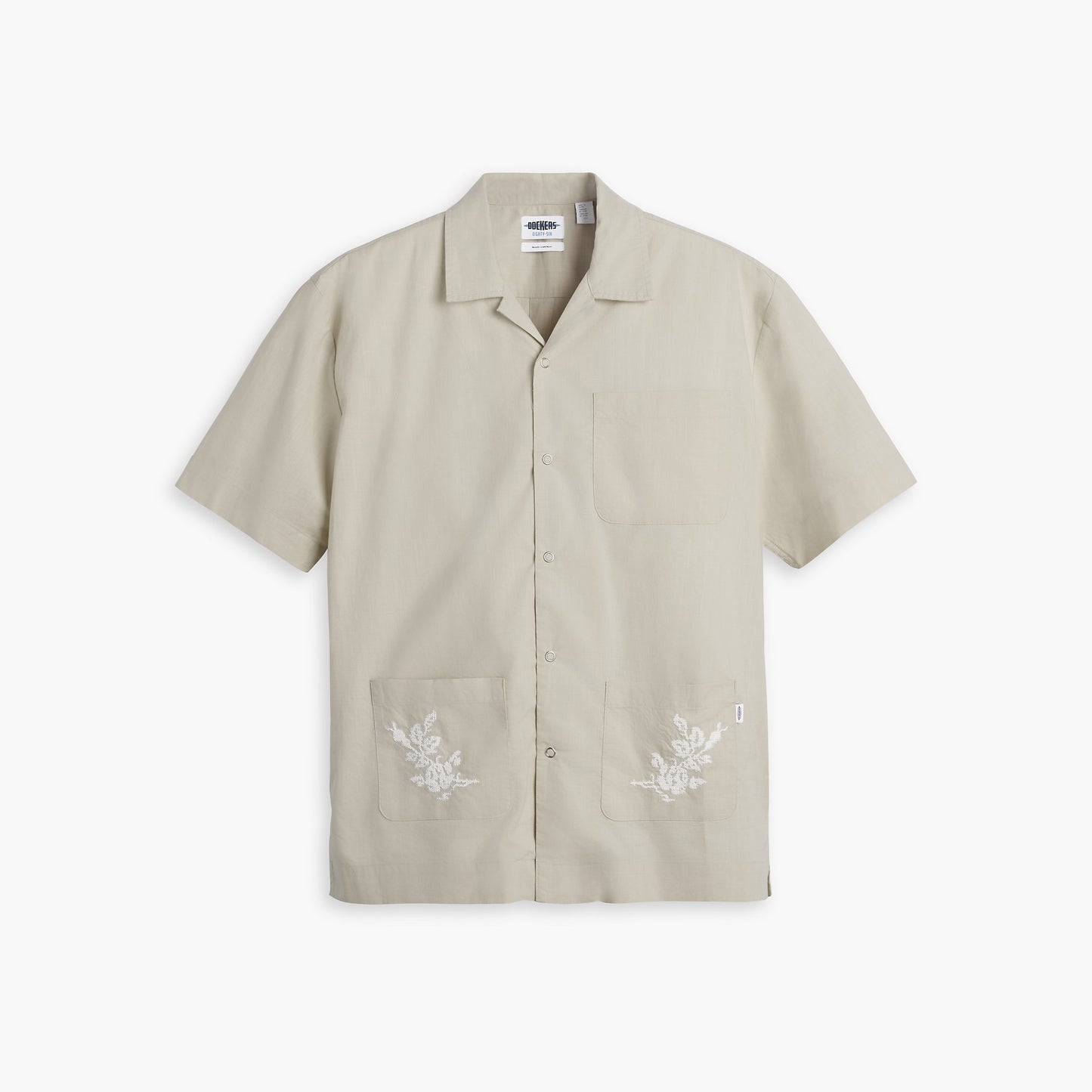 The Dockers Genderless 86 3 Pocket Shirt, SS is a beige short-sleeve, button-up shirt featuring a camp collar and floral embroidery on the front pockets. Crafted from 100% cotton for a relaxed fit, it is displayed against a white background.