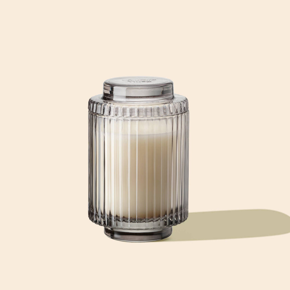 A cream-colored glass jar, featuring vertical ridges, contains La Jolie Muse's Amelie- Linen Cotton Oasis 7oz Candle, made from natural soy wax.