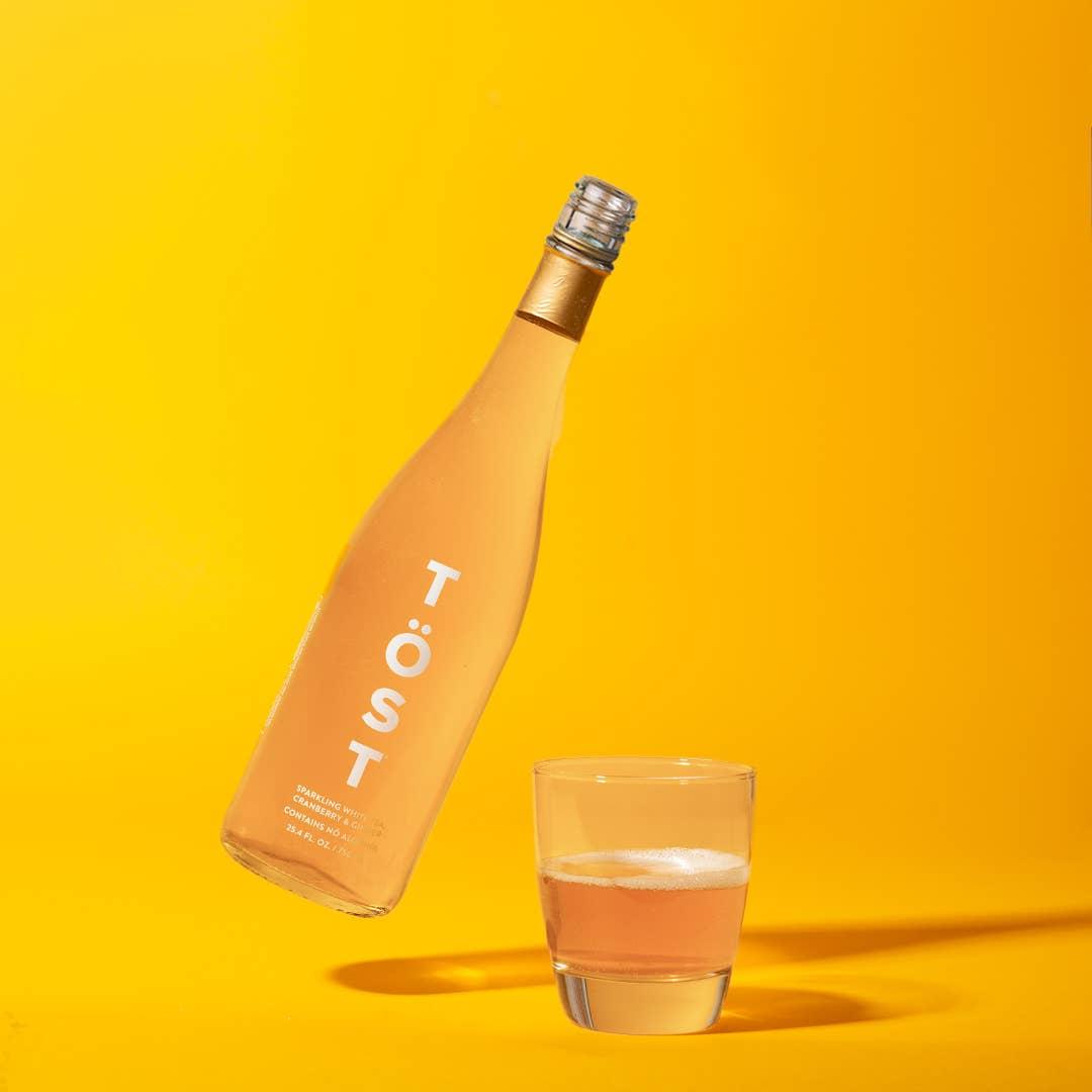 A bottle of TÖST, a non-alcoholic refresher, tilted at an angle with its cap off, next to a glass half-filled with the drink, both on a vivid yellow background.