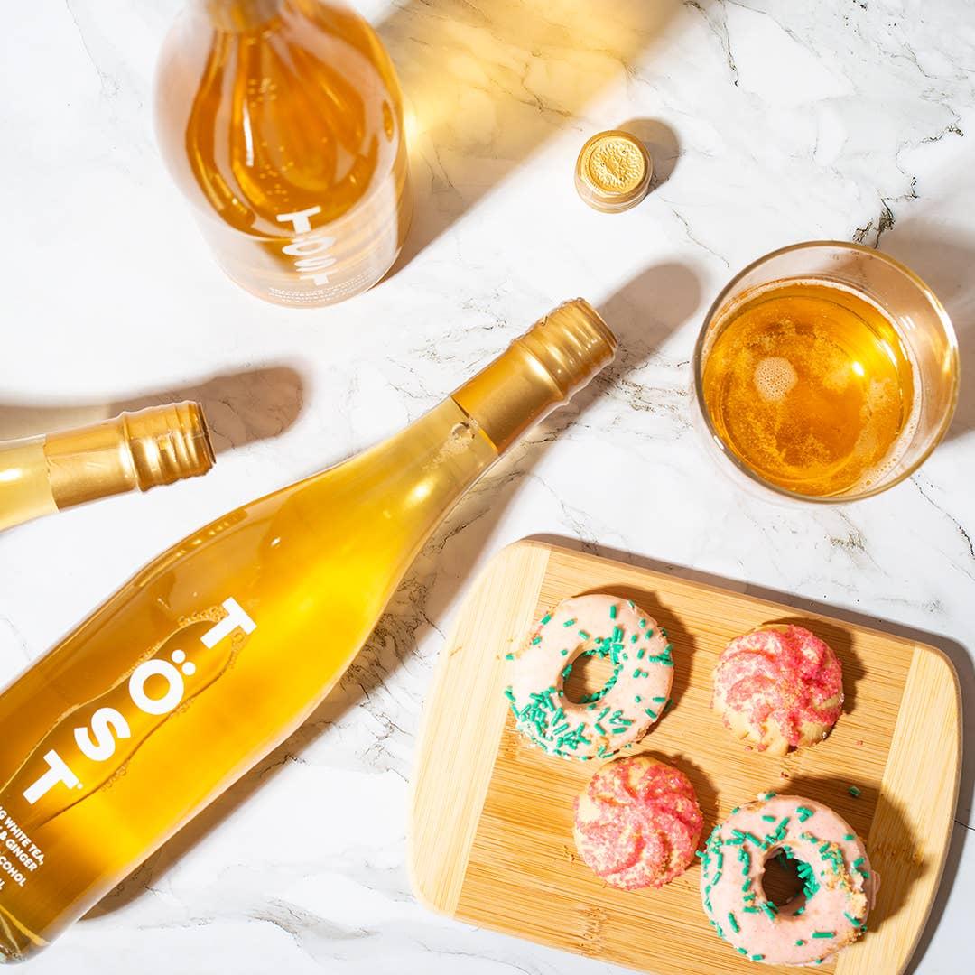 Two bottles of TÖST a Non Alcoholic Refresher and a glass filled with white tea, alongside a wooden tray of donuts with pink and green icing on a marble surface.