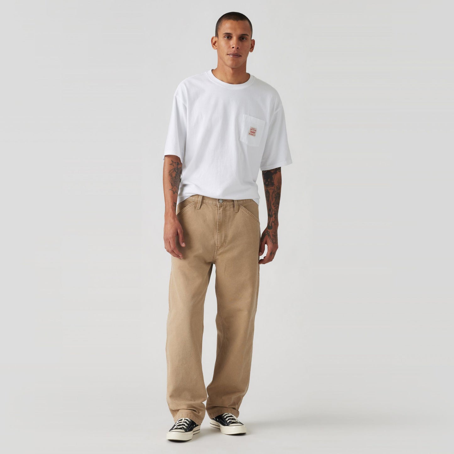 Against a plain backdrop, an individual in a neutral pose is wearing a white T-shirt, beige 568 Loose STRT Carpenter pants from Levis, and black sneakers.