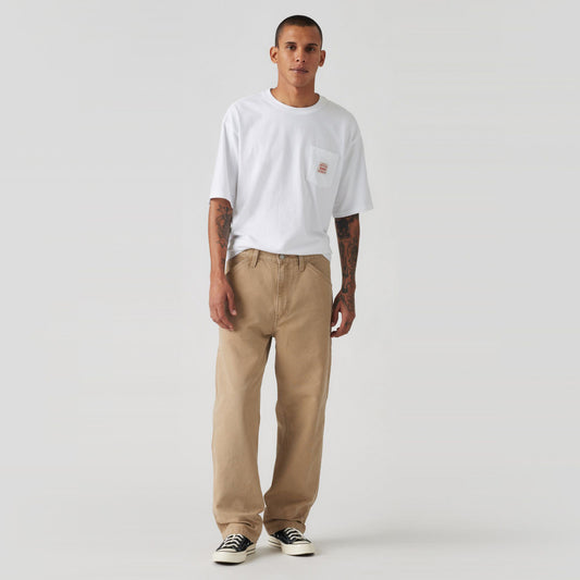 Against a plain backdrop, an individual in a neutral pose is wearing a white T-shirt, beige 568 Loose STRT Carpenter pants from Levis, and black sneakers.