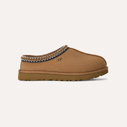 The W's Tasman mule slipper by Ugg features a tan suede upper, slip-on design, and brown rubber sole. It has decorative stitching around the ankle, the UGG logo on the side, and offers ultra-lightweight comfort with its cozy UGGplush™ lining.