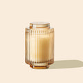 The Amelie - Tahitian Lychee 7oz Candle by La Jolie Muse, with its natural soy wax in a ribbed glass holder, exudes a touch of tropical sweetness against a simple backdrop.