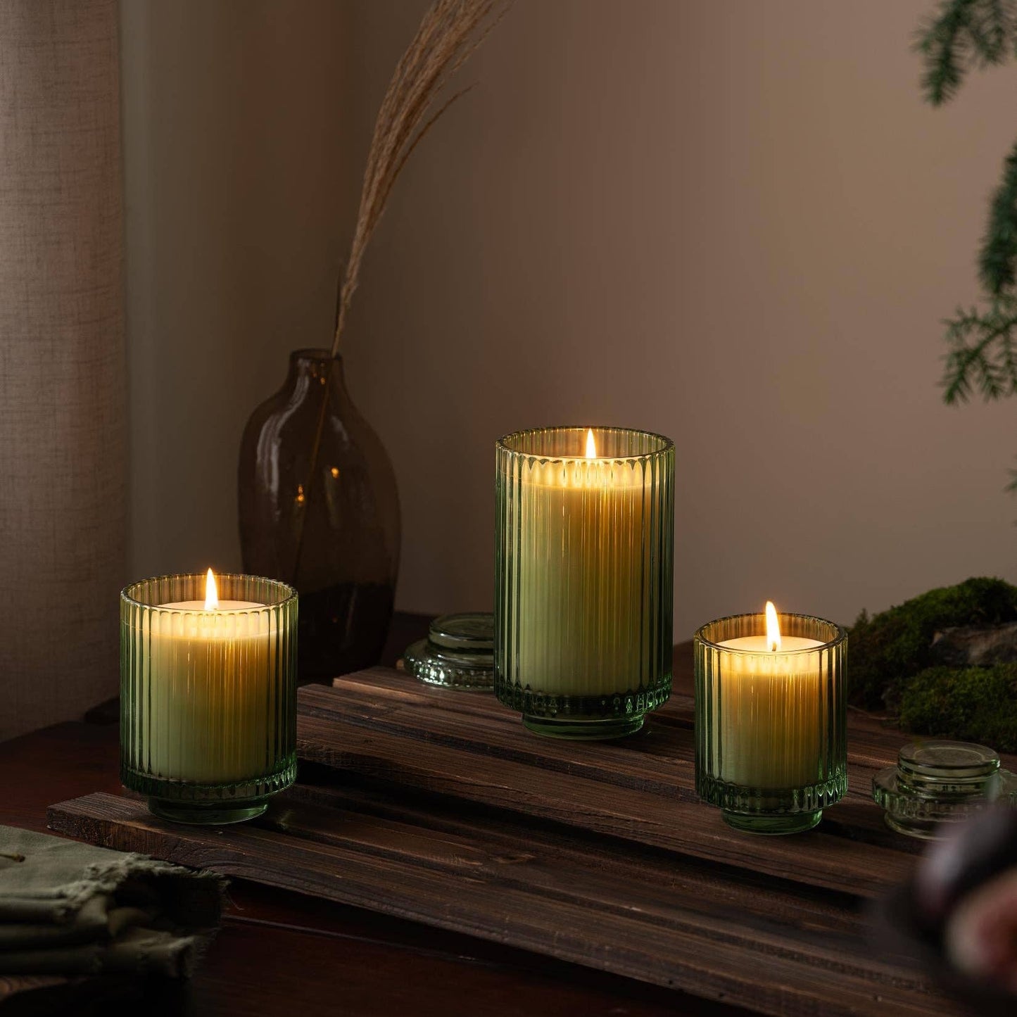 A trio of Amelie - Kyoto Vine & Moss 7oz candles by La Jolie Muse, housed in green ribbed glass holders and made from natural soy wax, placed on a wooden surface with decorative vases and greenery in the background.