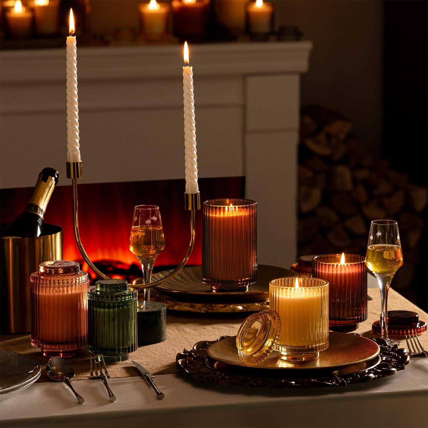 A cozy table setting adorned with ribbed glass candles, wine glasses, and decorative jars in front of a glowing fireplace is beautifully enhanced by the subtle fragrance of La Jolie Muse's Amelie - Tomato & Vine 7oz Candle.