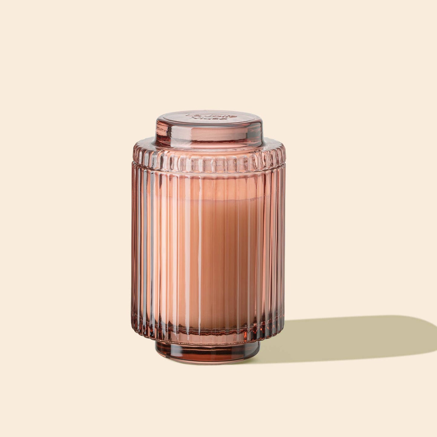 The Amelie - Watermelon Mint Zest 7oz Candle by La Jolie Muse, housed in a pink ribbed glass jar filled with natural soy wax, sits elegantly against a light beige background, casting a shadow to the right.