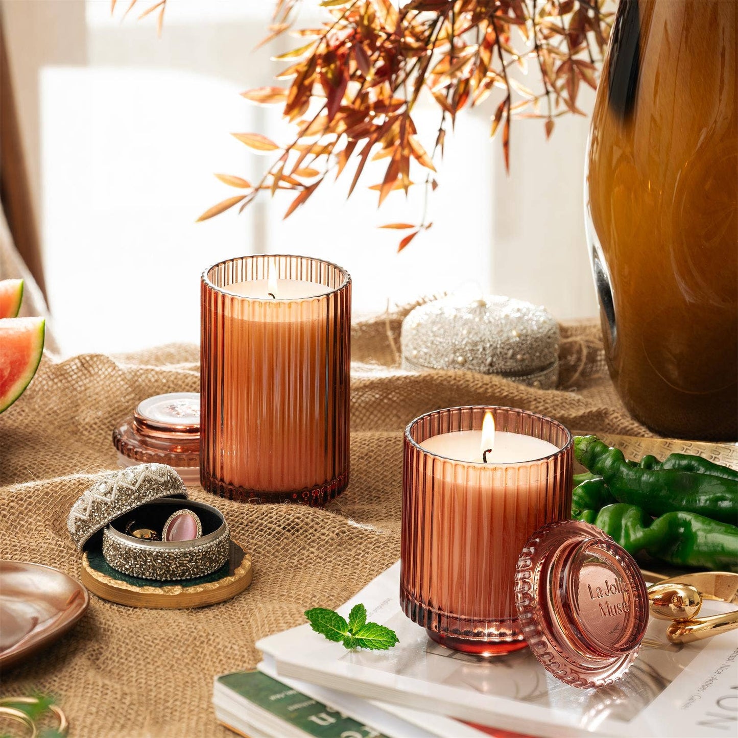 Two Amelie candles in a ribbed orange design, made from natural soy wax by La Jolie Muse, sit on a burlap-covered surface. They are surrounded by a plant, a small decorative box, and green peppers. Sunlight filters through a window in the background, enhancing the cozy ambiance with their watermelon mint zest scent.