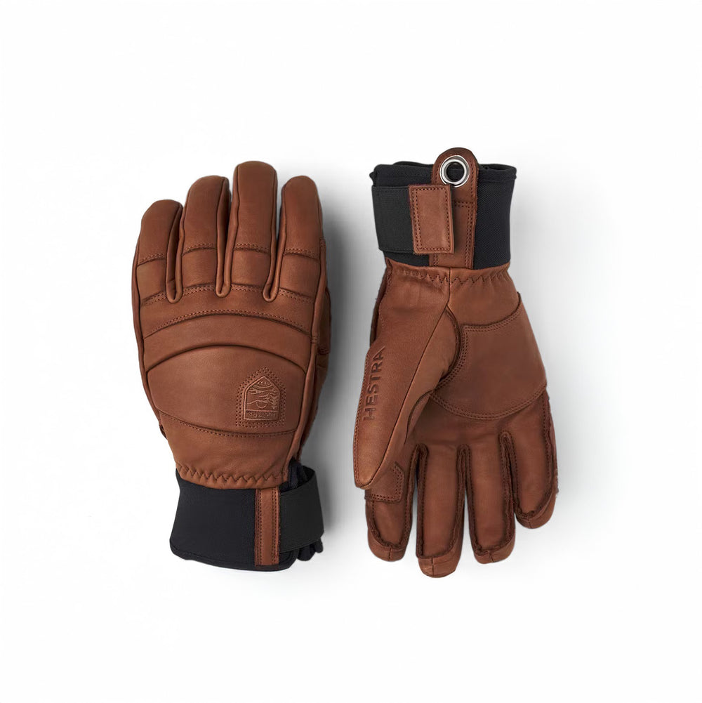 Two Fall Line Gloves in brown by Hestra, made from premium cowhide leather with black wrist cuffs featuring brand embossing and reinforced stitching. One glove is shown palm up and the other palm down against a plain white background.