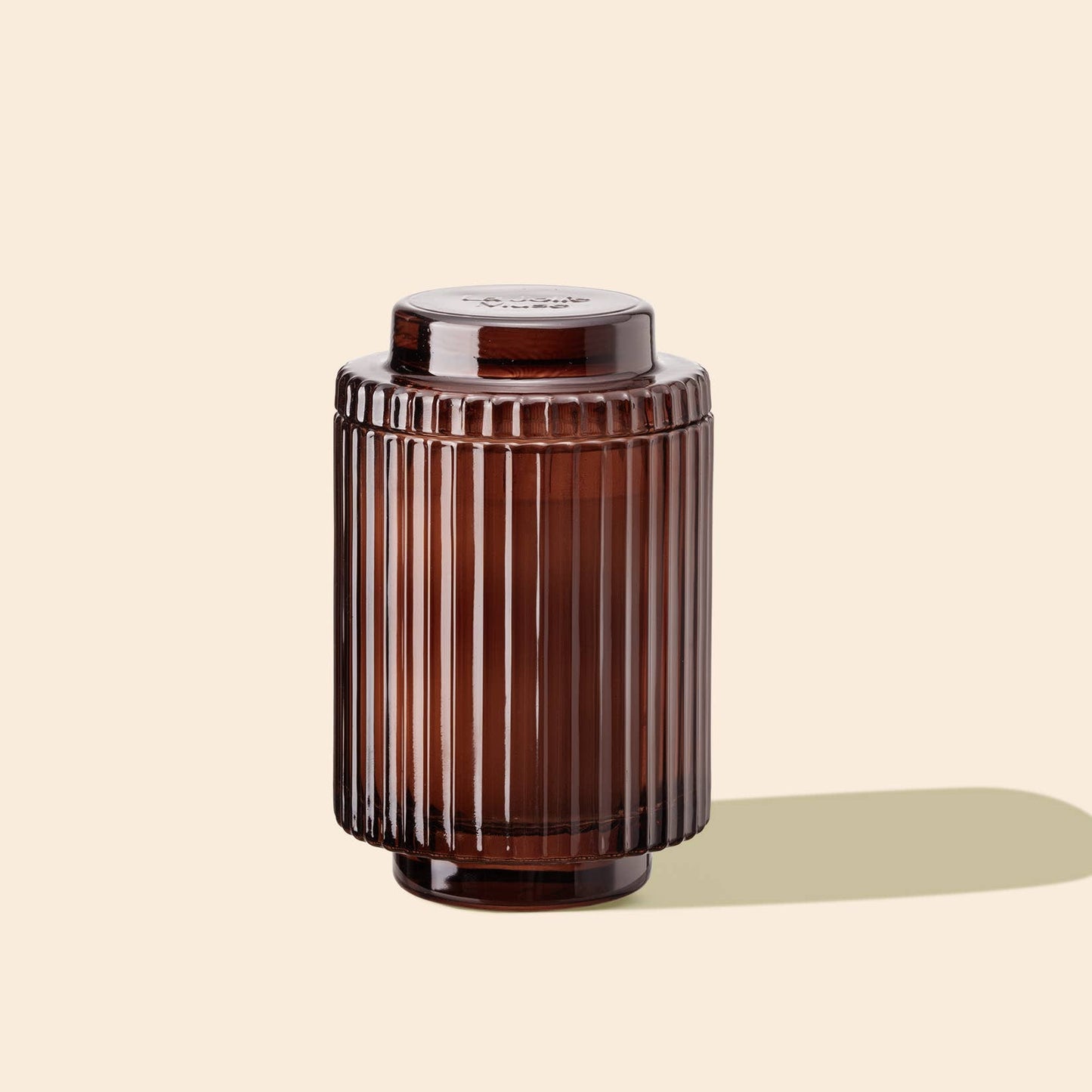 The Amelie - Tomato & Vine 7oz Candle by La Jolie Muse, featuring a brown ribbed glass jar with a lid, casts a shadow on a light beige background, evoking the look of ribbed glass candles.