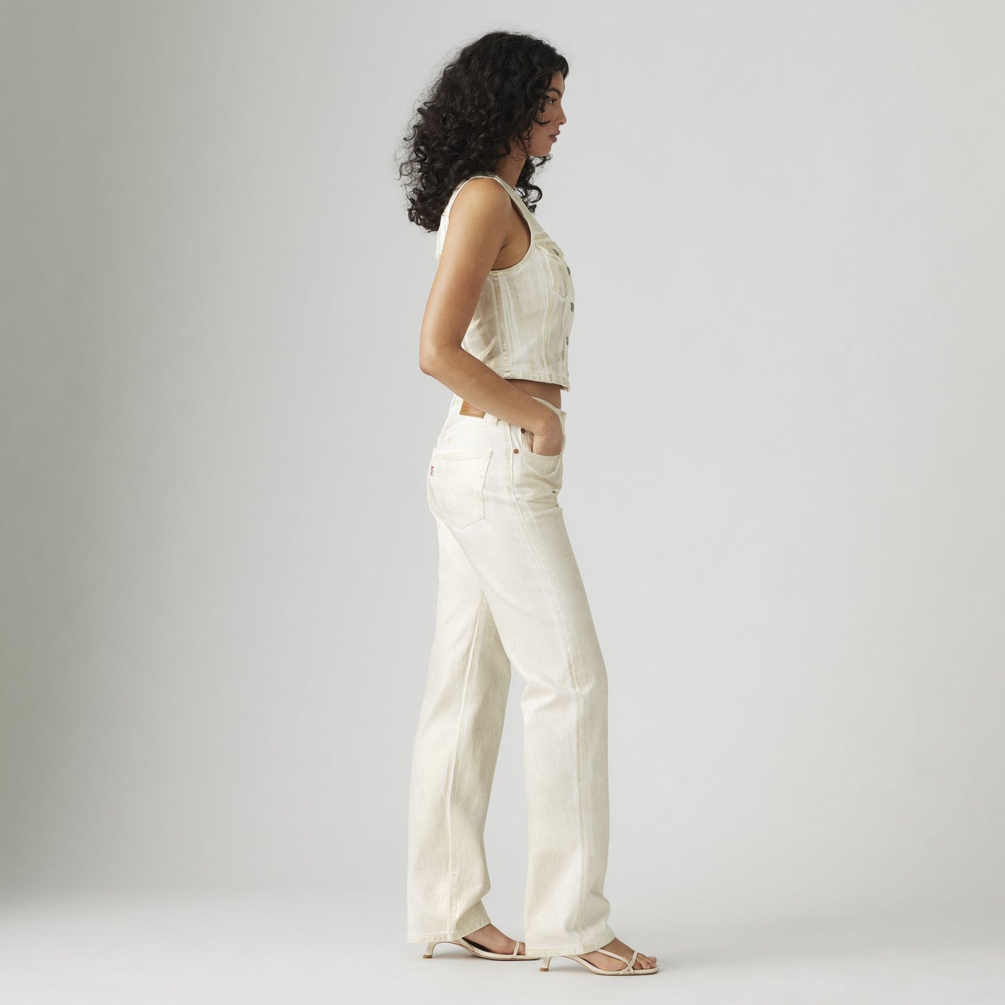 A woman with curly hair stands in profile against a plain background, wearing a light-colored sleeveless top, vintage Levis 501 90s jeans, and sandals.