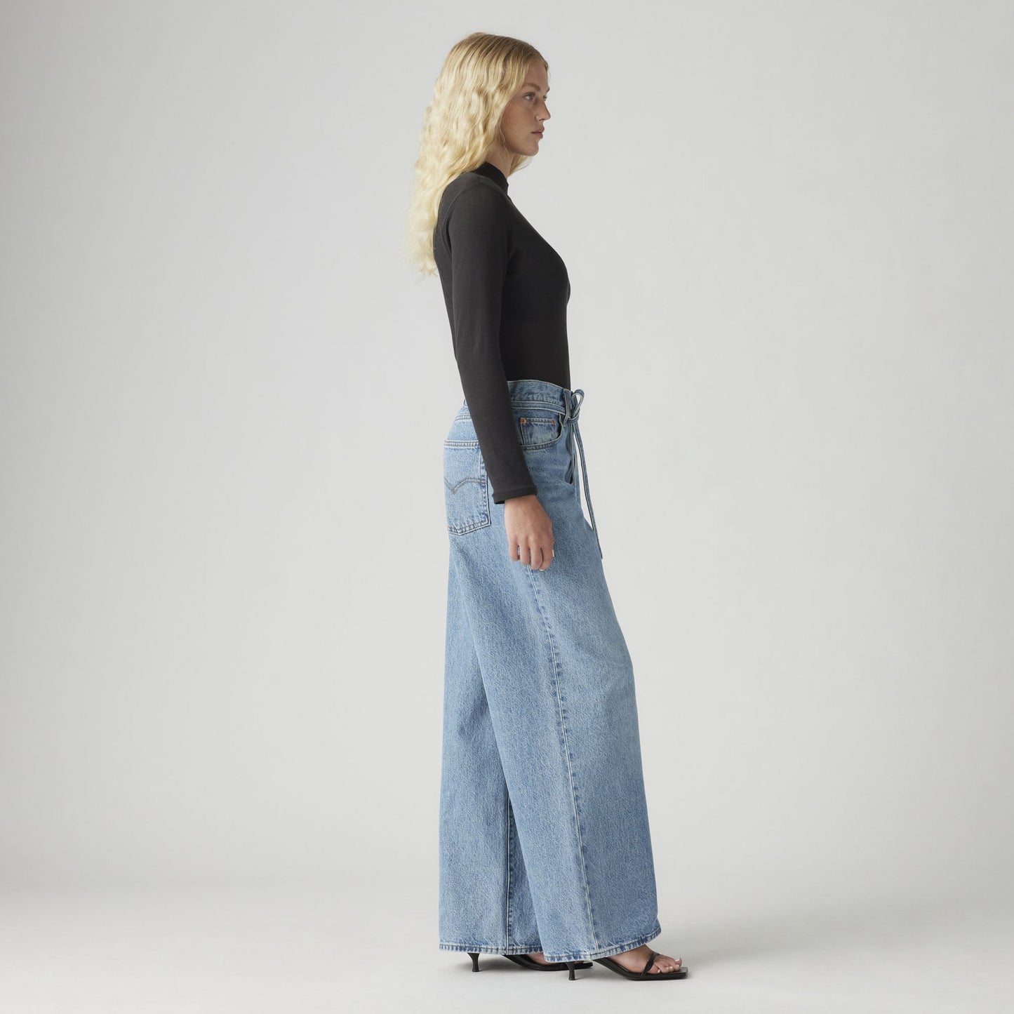 A person with long blonde hair is wearing a black long-sleeve top and Levis XL Straight oversized jeans, standing in profile against a plain background with heels.