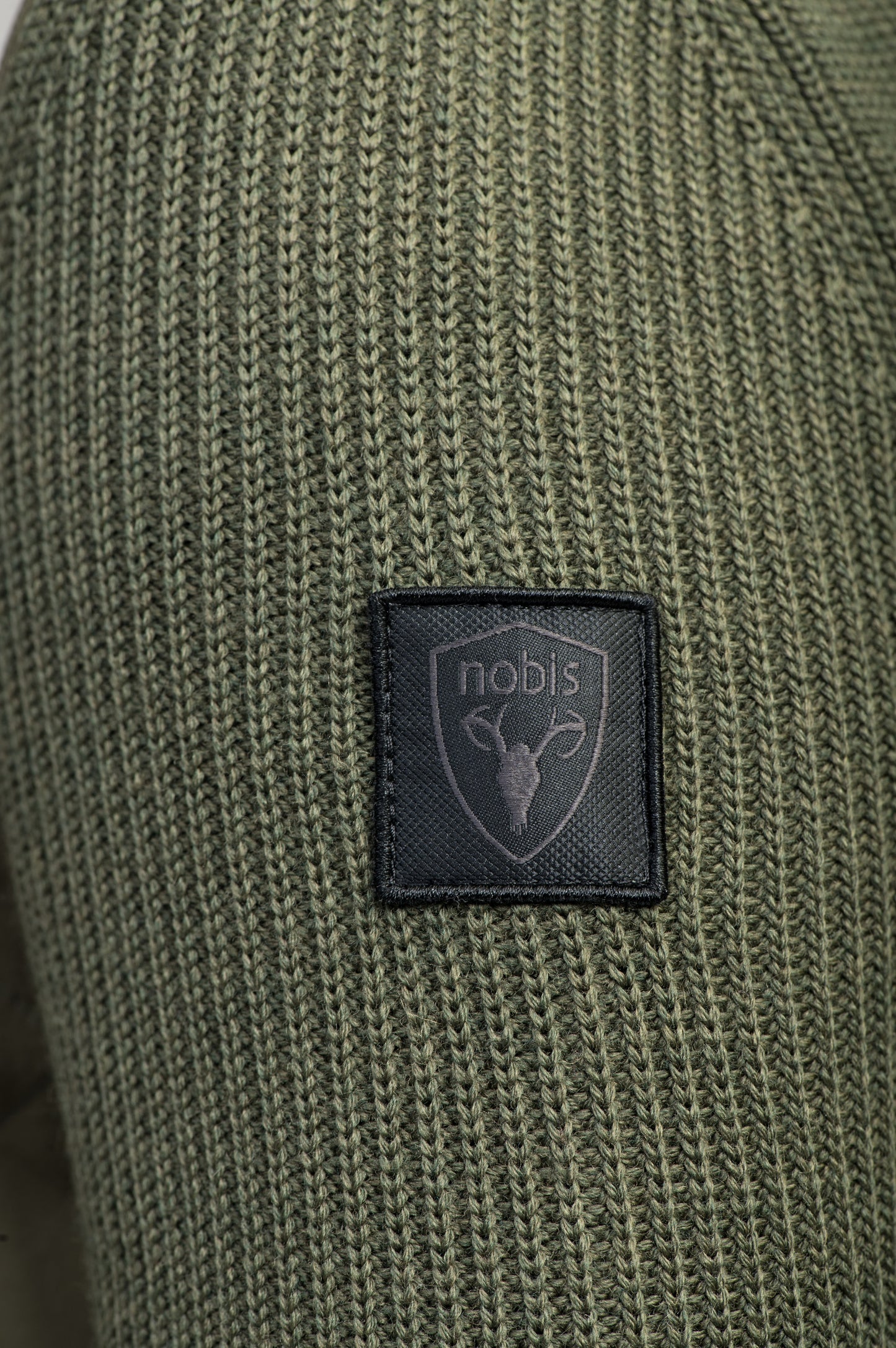 Close-up of Ada Women's Quilted Full Zip Sweater in green knit fabric featuring a black patch with a stylized deer head and "nobis" logo. This Nobis sweater offers warmth with Primaloft Gold Insulation Active+ and remains breathable.