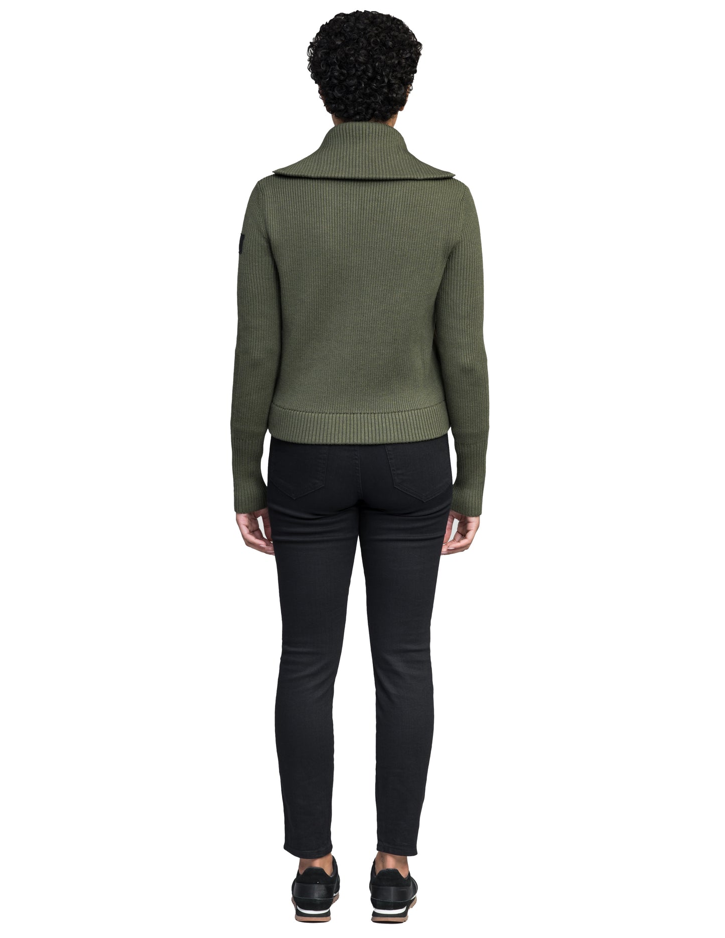 A person with short curly hair, facing away from the camera, wears a Nobis Ada Women's Quilted Full Zip Sweater in olive green and black pants.