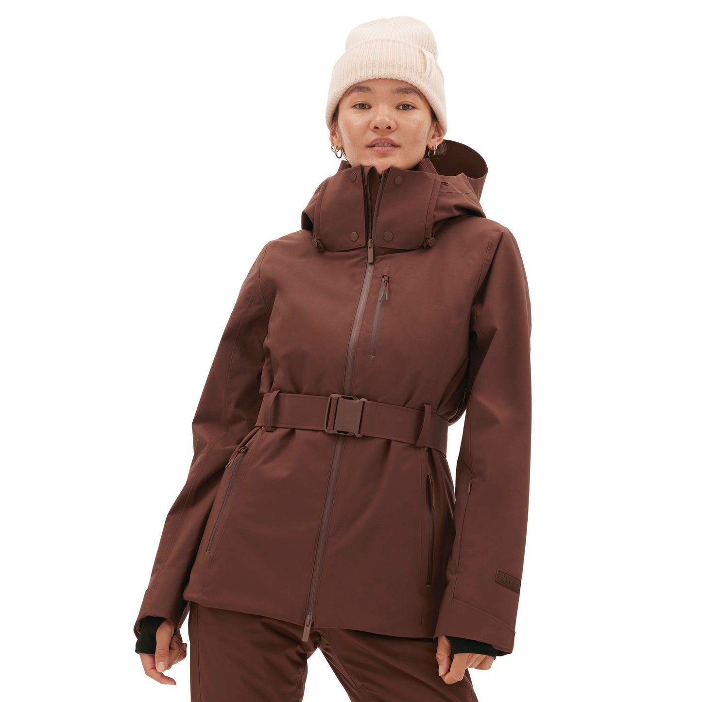 A person wearing the Aston Jacket from Halfdays, a brown belted ski jacket, and a white beanie stands against a white background.